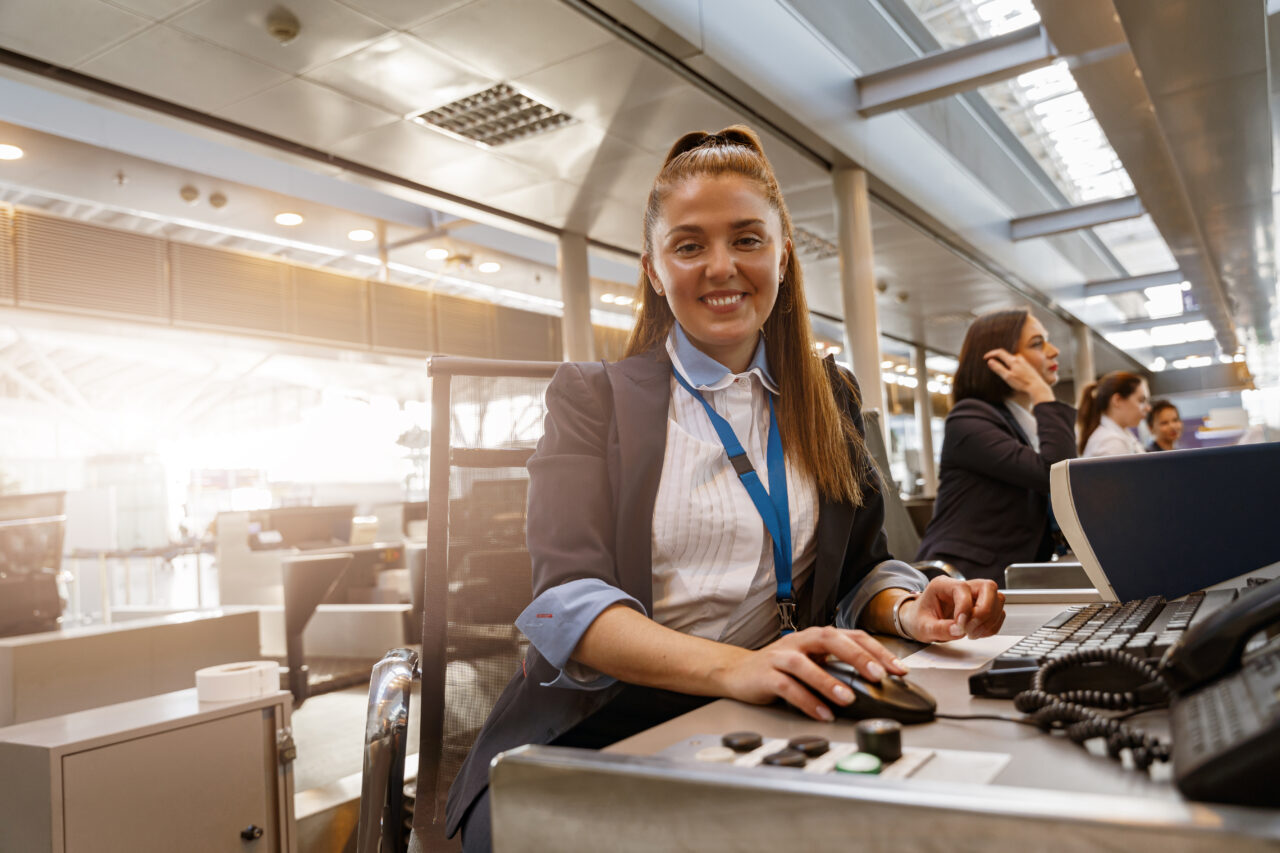 Woman airline employee working at airline check in counter in airport with colleagues on background